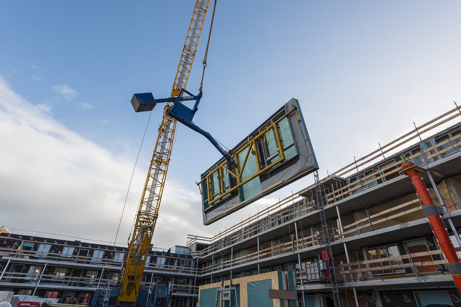 Een houtskeletbouwelement met geïntegreerde kozijnen hangt aan de kraan boven een woningbouwproject in aanbouw.