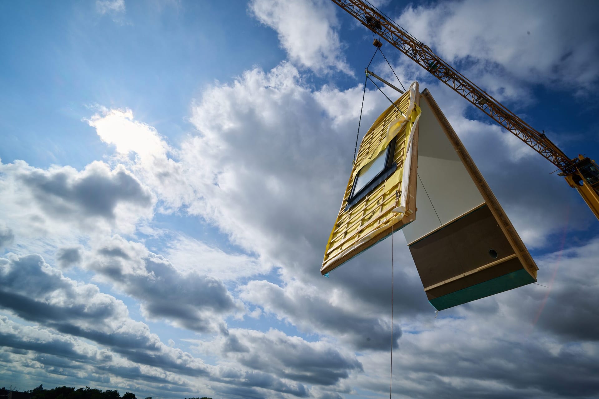 Een prefab dakelement met geïntegreerd dakraam hangt aan de bouwkraan tegen een wolkenlucht.