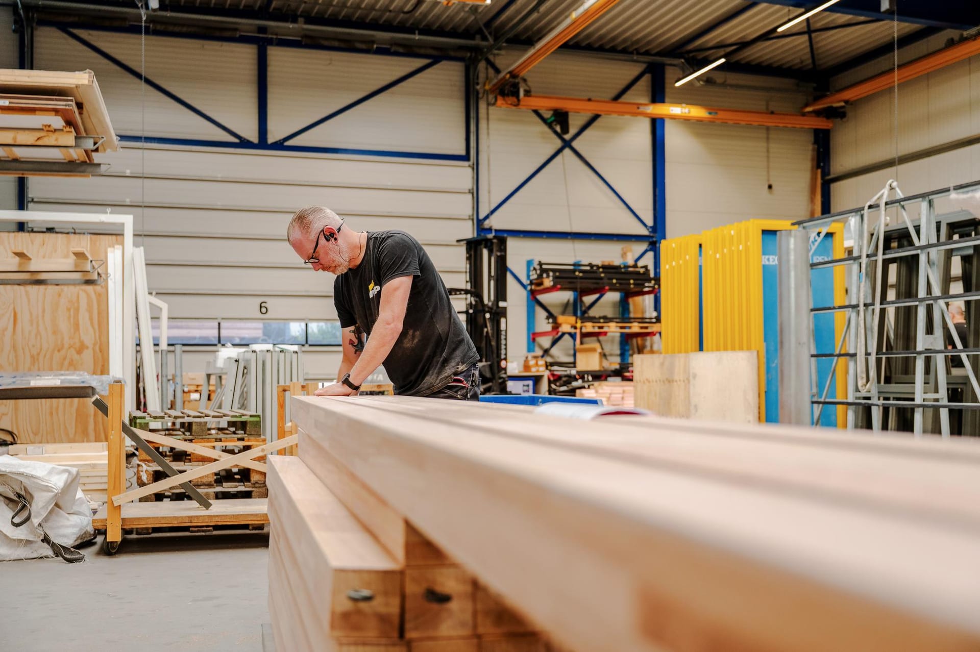 Medewerker controleert een pakket gelamineerde houten balken in de productiehal, met houten kozijnen op de achtergrond.