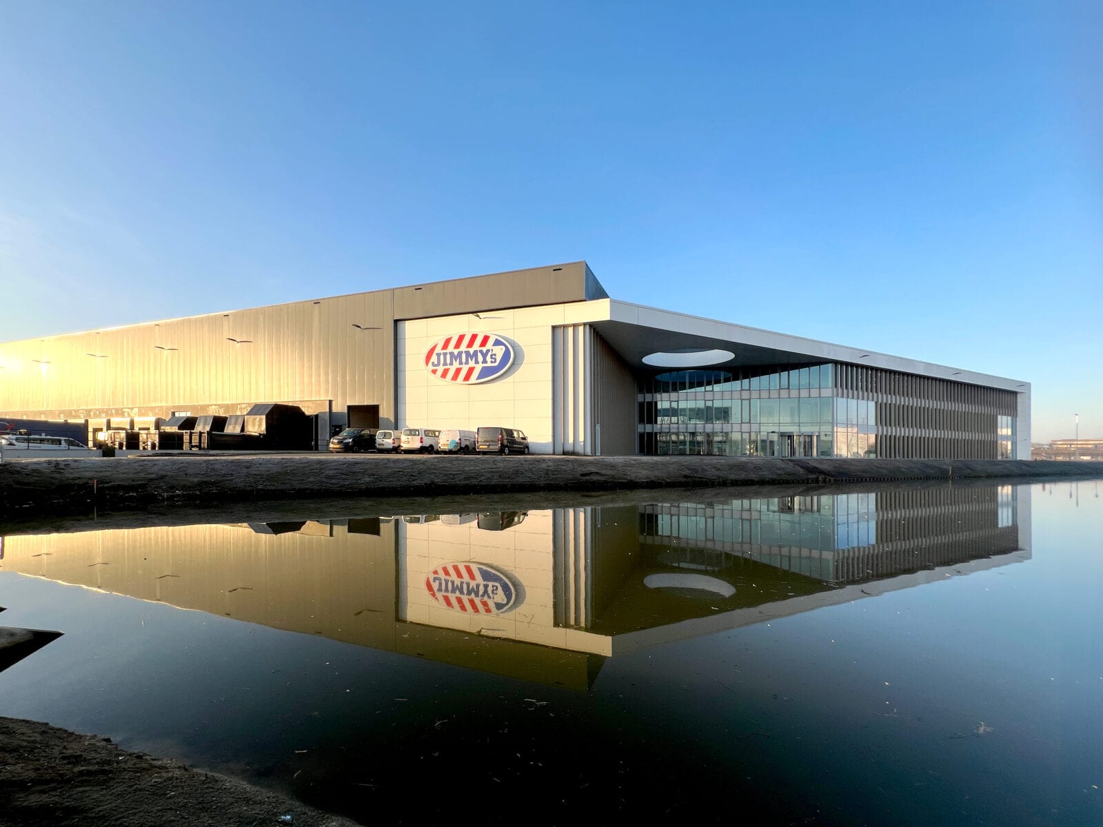 De hoofdvestiging van Jimmy Products in Ridderkerk met langgerekte gevel en glazen entreepartij, weerspiegeld in het water.
